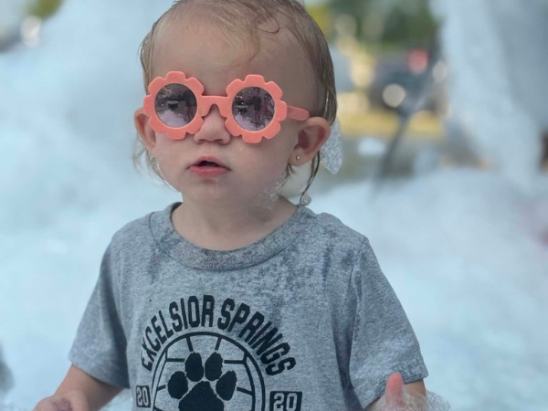 A young child in sunglasses plays in bubbles at a park.