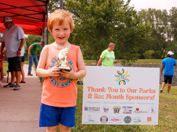 A child with a fish trophy stands next to a sign thanking Park and Recreation Month sponsors.