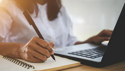 person writing with pen in notepad while using laptop
